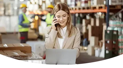 A smiling female manager in a business suit is talking on a phone and working on a laptop on top of wrapped pallets in a busy warehouse.